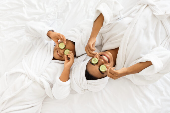 Beauty Spa Day. Cheerful Mom And Daughter In Bathrobes Lying With Cucumber Slices On Eyes, Doing Face Mask Treatment, Wearing Towel On Head, Having Fun Together At Home, Top View